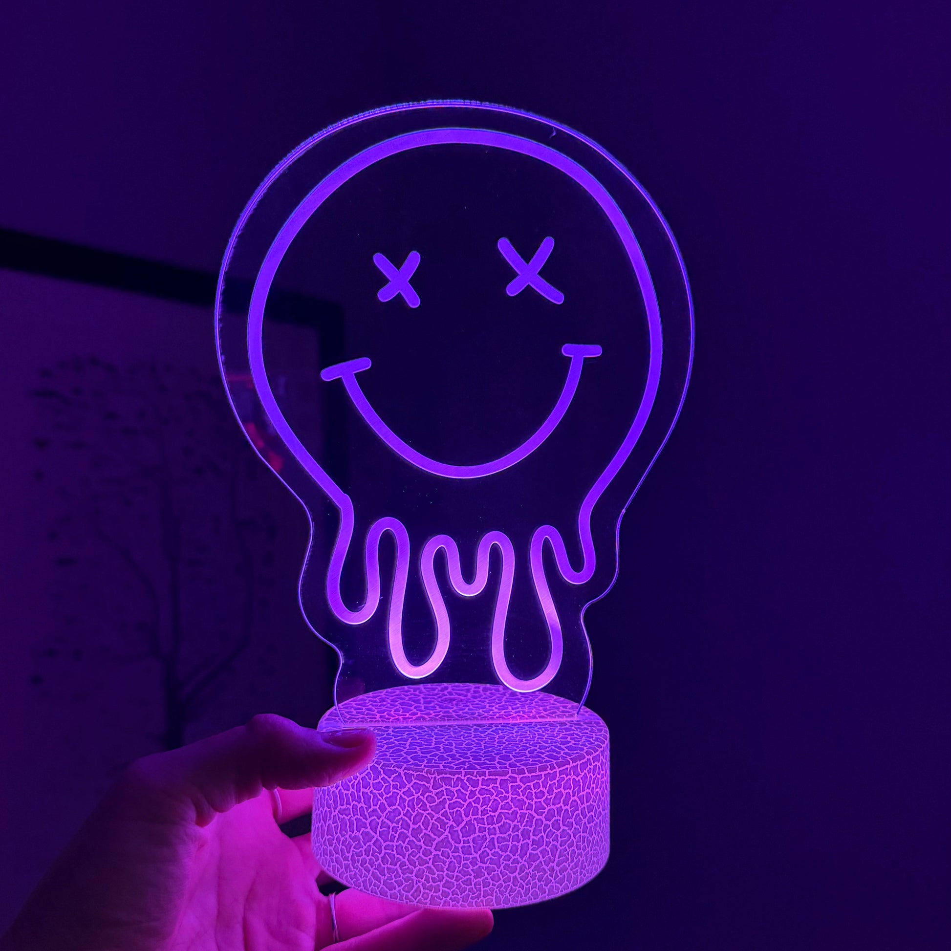 Neon light shaped like a smiley face with crossed eyes, held in a hand against a dark background.
